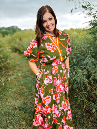 Woman in a floral dress standing in a field. Dress is olive green midi with pink and red florals, puffed sleeves, and contrasting stripes on the v neckline. Made by THML