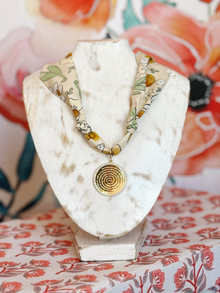 flay lay style photo with a Taupe scarf with greens and yellow floral print with a gold plated pendant featuring the Lord's Prayer in a spiral pattern and encrusted in crystals along the outer edge
