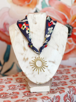 flat lay style image of a navy scarf necklace with bold floral print and gold plated textured sunburst pendant