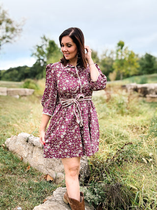 woman wearing plum colored floral mini dress with unique crochet trim, self tie sash, and button down placket