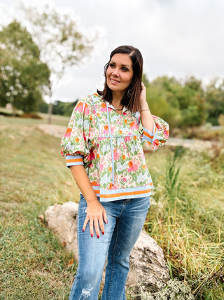 Woman wearing a floral blouse and jeans standing in a natural setting with trees and grass. Her top is Blousy Oversize Top with bold Floral Print and Light Blue and Orange Striped hem and cuffs and neck tie strings.