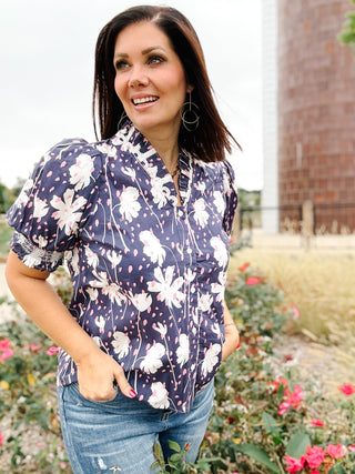 Woman wearing a blue floral button down blouse with ruffle neckline standing outdoors with flowers and a building in the background