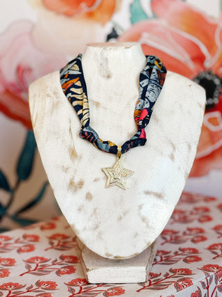 Flat lay style photo of a large gold plated star pendant scarf necklace on a navy scarf with bright pops of color in an abstract print