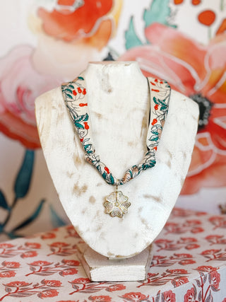 Flat Lay display of a gold plated clover pendant covered in crystals on a scar necklace in cream background, corals and green florals