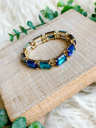 Bracelet with blue and green gemstones on a wooden block with green leaves in the background