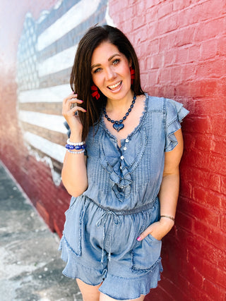 Woman wearing a washed denim ruffle romper with flutter sleeves, drawstring waist, front pockets, and feminine ruffle details, perfect for Fourth of July celebrations, summer BBQs, concerts, and casual Americana style.