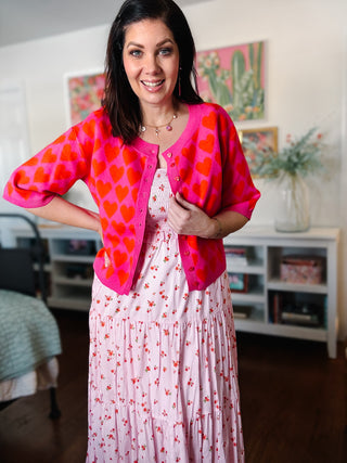 Woman wearing a pink heart-patterned cardigan over a white dress in a room with colorful artwork on the wall.