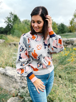 Woman wearing a colorful sweater with abstract patterns in an outdoor setting. About her top: Grey Puffed Sleeve Luxe Sweater with Rust Navy Pink and Cream Abstract Floral Print and Striped Neckline and Cuffs by THML