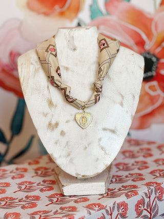 flat lay style image of a Taupe scarf necklace with geometric pink and brown diamond pattern and a gold plated heart pendant featuring Mary