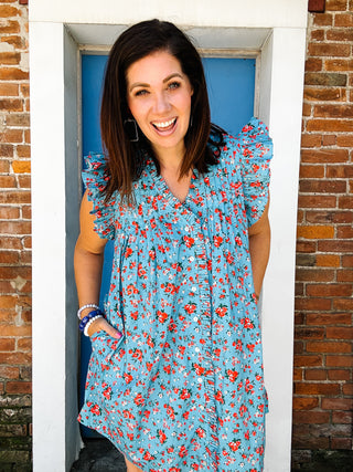 Woman wearing a blue floral mini dress with ruffle sleeves, front button details, and pockets.