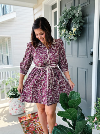 A woman wearing a short-sleeve button-front dress with a purple and white floral pattern and a matching tie belt, standing on a front porch in front of a gray door with a green wreath.