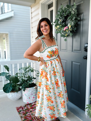 Woman wearing the Cecelia Maxi Dress featuring a cream background with orange floral print, bow-tie shoulder straps, square neckline, smocked back, pockets, and a flowy maxi skirt.