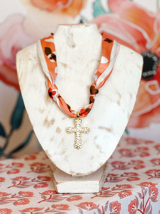 flat lay style photo with a coral scarf floral necklace with gold plated white cross pendant with scroll detailing