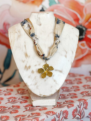 Flay lay style photo of a cream twilly scarf with navy and white floral print with a large textured 5 petal flower gold plated pendant