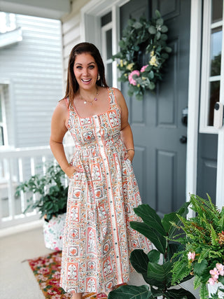 Woman wearing the Amalfi Midi Dress featuring a vintage floral tile print, coral scalloped trim, square neckline, button front, smocked back, and hidden pockets.