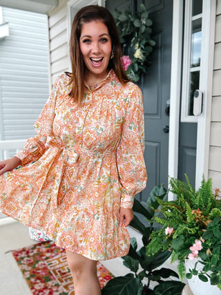 Woman wearing the Paris Mini Dress featuring a vintage floral print, long sleeves, stand collar, adjustable waist tie, tiered skirt, and hidden side pockets.