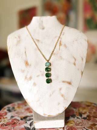 Gold-tone necklace with four vertically arranged round faceted stones, featuring one aqua stone above three emerald green stones, displayed on an ivory jewelry bust.