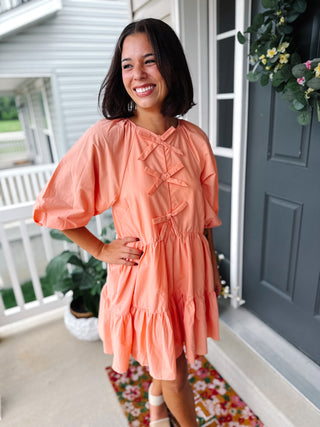 Woman wearing the Karlie Bow Poplin Dress in Papaya featuring statement front bows, puff sleeves, lightweight cotton poplin fabric, hidden pockets, and a relaxed tiered mini dress silhouette.