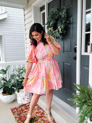 Woman wearing a pink floral shirt dress by Karlie with a matching belt, puff sleeves, button front, and tiered skirt.