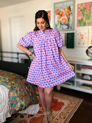 Woman wearing a lavender dress with blue patterns in a room with floral decor.