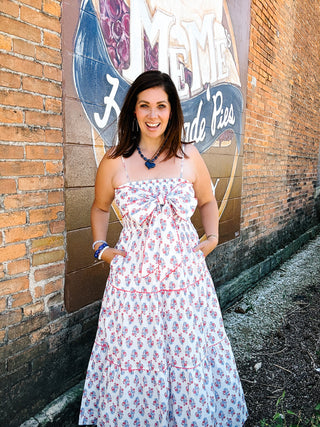 Woman wearing an ivory floral maxi dress with red and blue floral print, oversized bow bodice, adjustable straps, and tiered skirt.