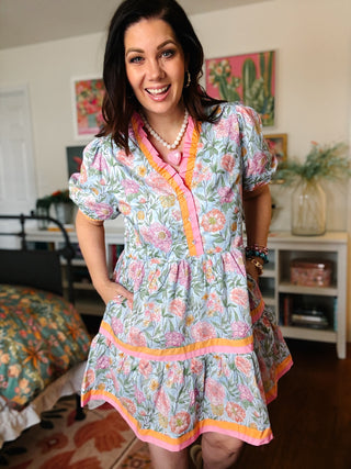 Woman wearing a colorful floral boutique mini dress with puff sleeves, contrast pink and orange trim, tiered skirt and pockets.