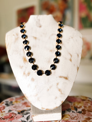 Navy blue faceted stone statement necklace featuring round dark blue stones in gold-tone prong settings displayed on a cream jewelry bust.