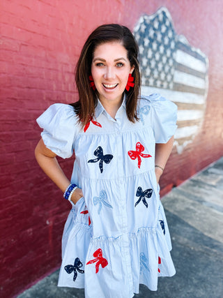 Woman wearing a light blue patriotic bow embroidered dress with puff sleeves and red, white, and blue bow details.