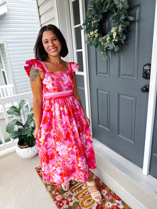 Woman wearing a pink floral empire waist midi dress featuring a square neckline, flutter sleeves, hidden side pockets, and a flowy midi-length skirt.