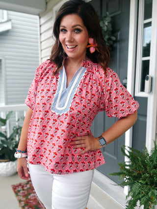 Woman wearing a pink floral puff sleeve blouse with a contrast blue striped scalloped neckline and white jeans.