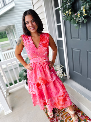 Woman wearing a pink floral wedding guest midi dress featuring embroidered scalloped trim, flattering side waist cutouts, a sleeveless bodice, and a tiered midi-length skirt.