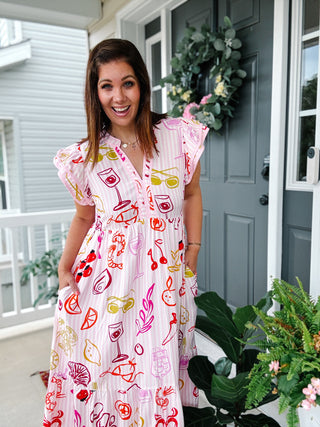 Woman wearing the Sardinia Midi Dress featuring pink stripes, colorful Mediterranean food illustrations, flutter sleeves, tiered midi skirt, and side pockets.