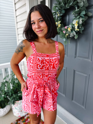 Woman wearing a red and pink printed romper with adjustable straps, smocked back, pockets, functional buttons, and a removable waist tie.