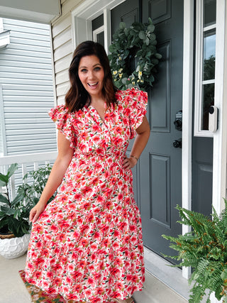 Woman wearing the THML Primrose Midi Dress featuring a vibrant pink coral floral print, flutter ruffle sleeves, a flattering smocked waist, hidden side pockets, and a flowy tiered midi silhouette.