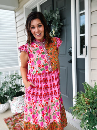 Woman wearing a pink floral patchwork midi dress featuring mixed botanical prints, flutter sleeves, a button-front bodice, hidden side pockets, and a flowy tiered cotton skirt by thml