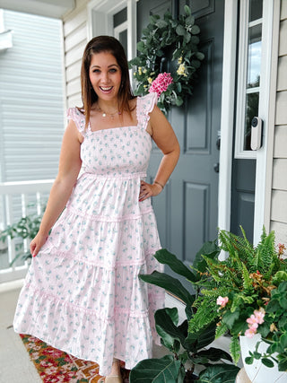 Woman wearing the Vienna Midi Dress featuring a blush pink floral print, smocked bodice, ruffle straps, hidden side pockets, and a flowy tiered midi silhouette.