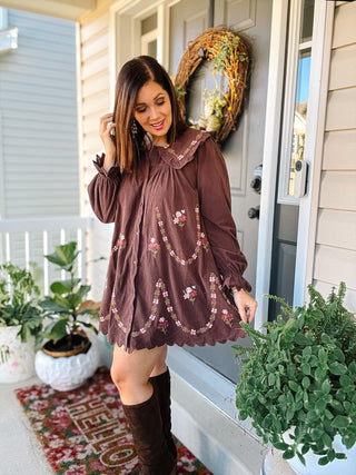 Woman wearing Winnie chocolate brown floral embroidered mini dress with ruffled collar, scalloped hem and tall brown boots.